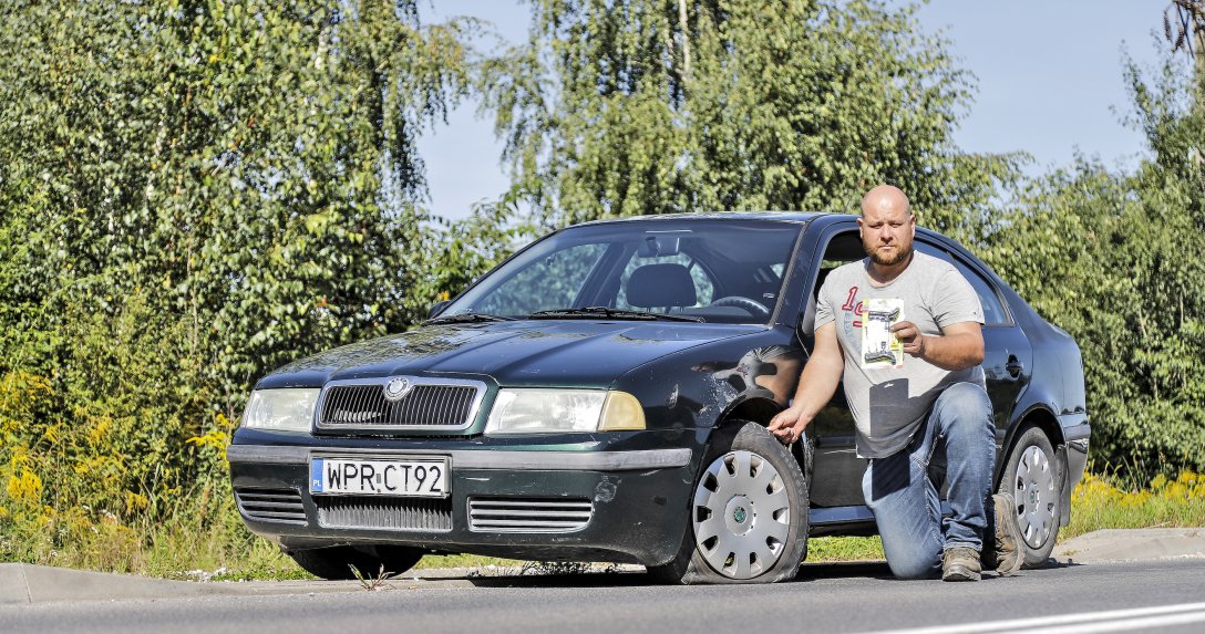 Człowiek przy aucie z przebitą oponą pokazuje sznur do uszczelniania